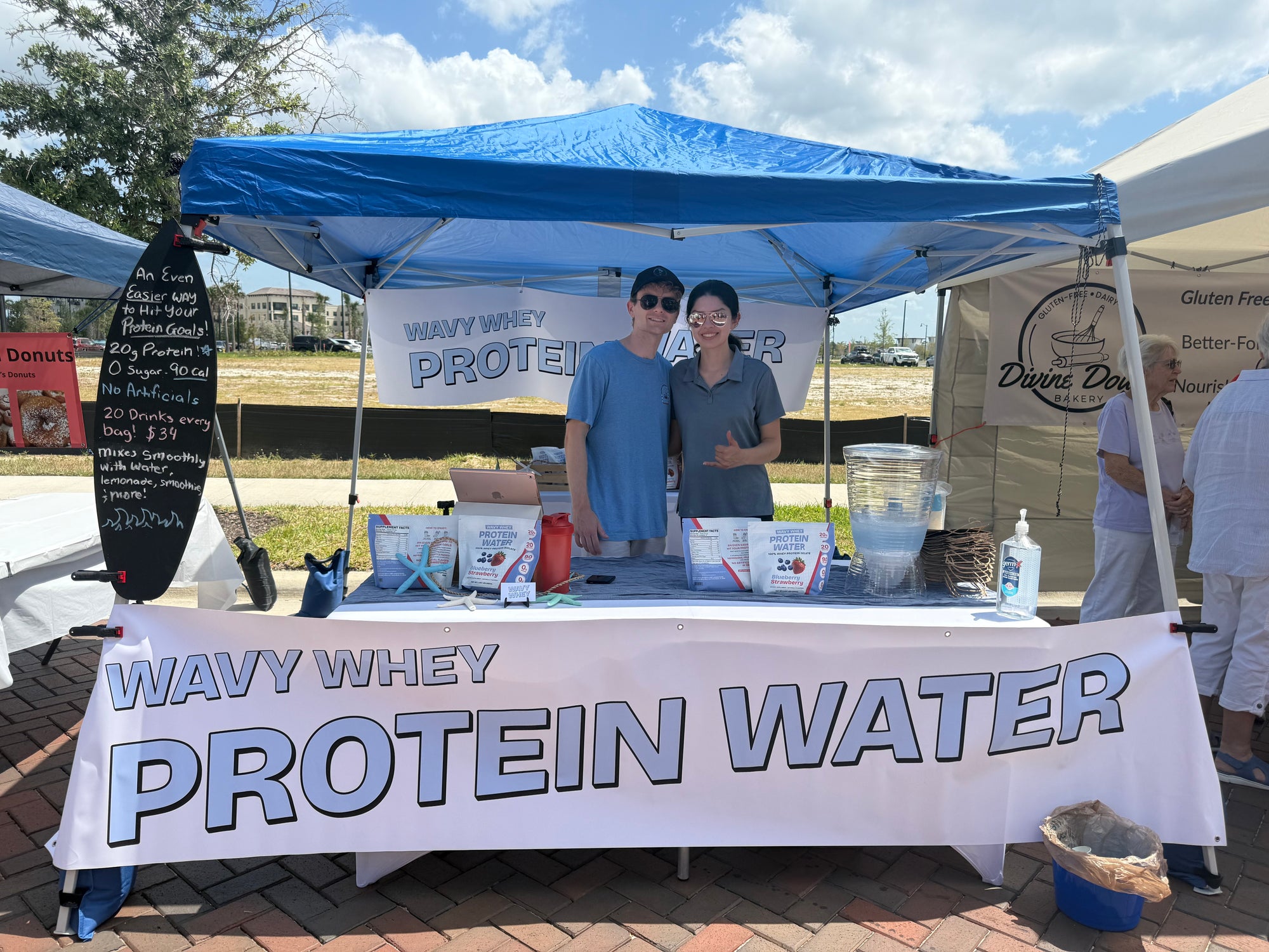 Two co-founders standing behind a 'Wavy Whey Protein Water' booth at Wellen Park Farmer's Market.