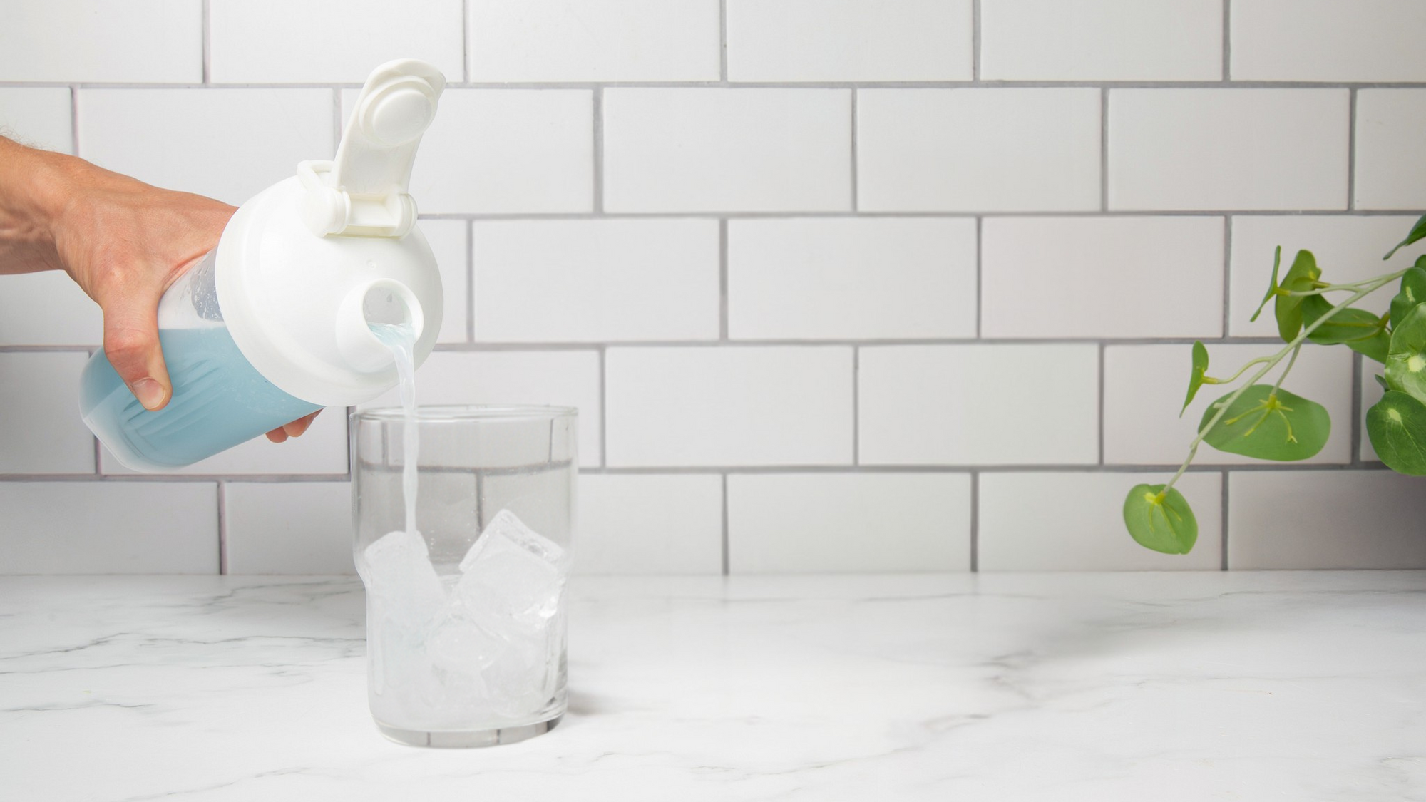 Person pouring a liquid from a bottle into a glass jar with a white tiled wall background