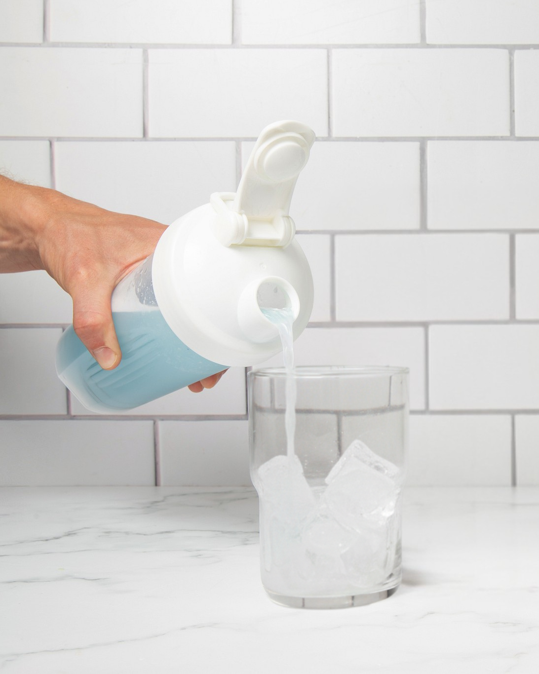 Person pouring a liquid from a blue and white container into a glass jar against a tiled wall background.